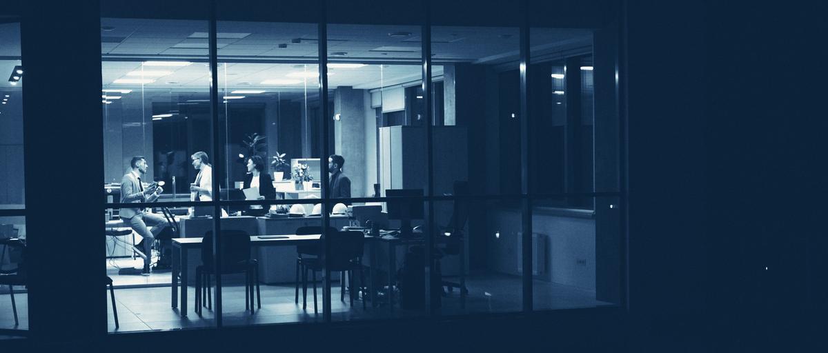 An office floor lit up in the evening, seen from outside: a team at work behind a large window.